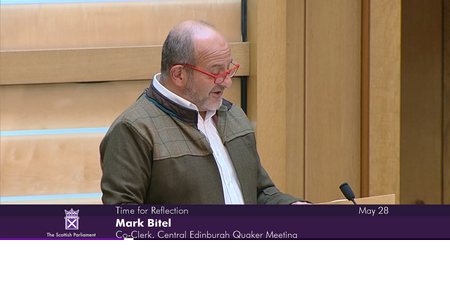 Mark Bitel, Co-Clerk of Central Edinburgh Quaker Meeting, addresses Scottish Parliament. Mark is wearing a white shirt, green jacket and red glasses.