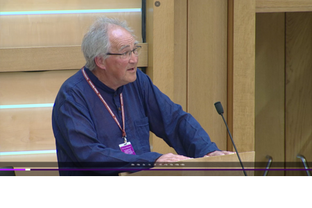 Richard Raggett, a Quaker from North Scotland, speaks to Parliament. Richard has grey hair, glasses, and is wearing a dark blue shirt standing in front of the podium in the Scottish Parliament