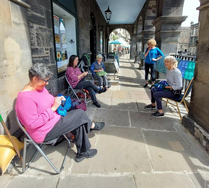 Loving Earth Project action in Edinburgh A socially distanced group of people sit outside a Meeting House. They are crafting embroidered panels. You can see more panels in the window of the Meeting House behind them. They look friendly and approachable.
