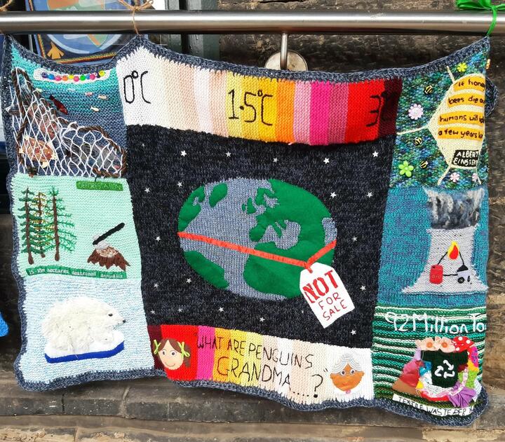Knitted panel, created by Quakers working with the Loving Earth Project. A knitted panel with an earth in the centre, circled by a ribbon and tag saying "not for sale". Various images associated with the climate crises border it.