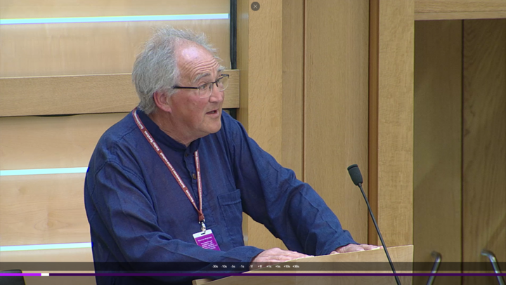 Richard Raggett, a Quaker from North Scotland, speaks to Parliament. Richard has grey hair, glasses, and is wearing a dark blue shirt standing in front of the podium in the Scottish Parliament