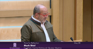 Mark Bitel, Co-Clerk of Central Edinburgh Quaker Meeting, addresses Scottish Parliament. Mark is wearing a white shirt, green jacket and red glasses.