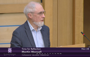Picture of Quaker Martin Mansell giving the Time for Reflection. He has grey hair, a beard, and glasses and is wearing a grey suit. He is in front of the lectern in the Debating Chamber of parliament.