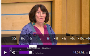 A woman stands before a microphone at the Scottish Parliament, delivering a speech